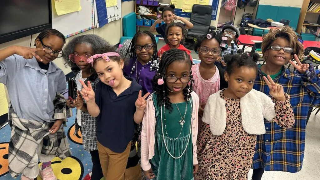 A group of approximately 12 elementary students poses in a classroom, with many dressed up as elderly people to celebrate the 100th day of school. They are smiling and making peace signs for the camera.
