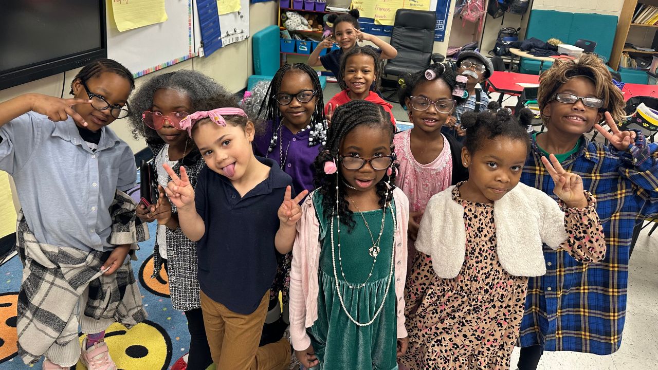 A group of approximately 12 elementary students poses in a classroom, with many dressed up as elderly people to celebrate the 100th day of school. They are smiling and making peace signs for the camera.