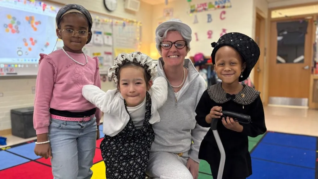 Three young students and one adult pose in a classroom while dressed as elderly people for the 100th day of school. The students are wearing costumes including a grey wig, a floral bonnet, and a black hat with a walker, while the adult wears a white wig and glasses.