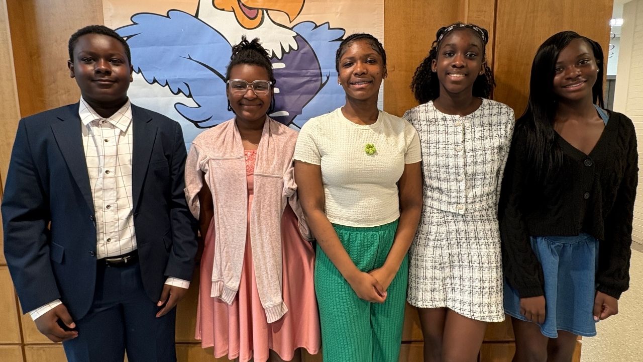 Five dressed-up students standing in a row and smiling in front of a wood-paneled wall featuring an eagle mascot banner.