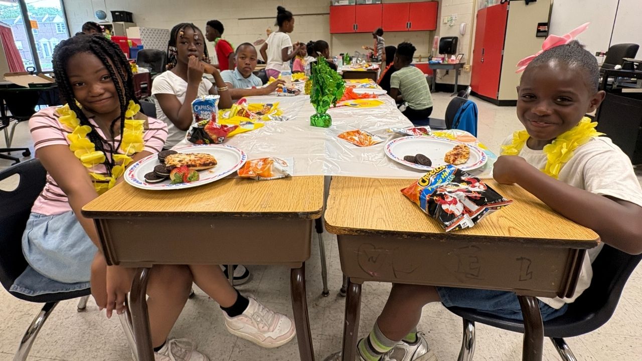 Students wearing yellow flower leis sitting around classroom desks gathered for a party with snacks, juice pouches, and pizza.