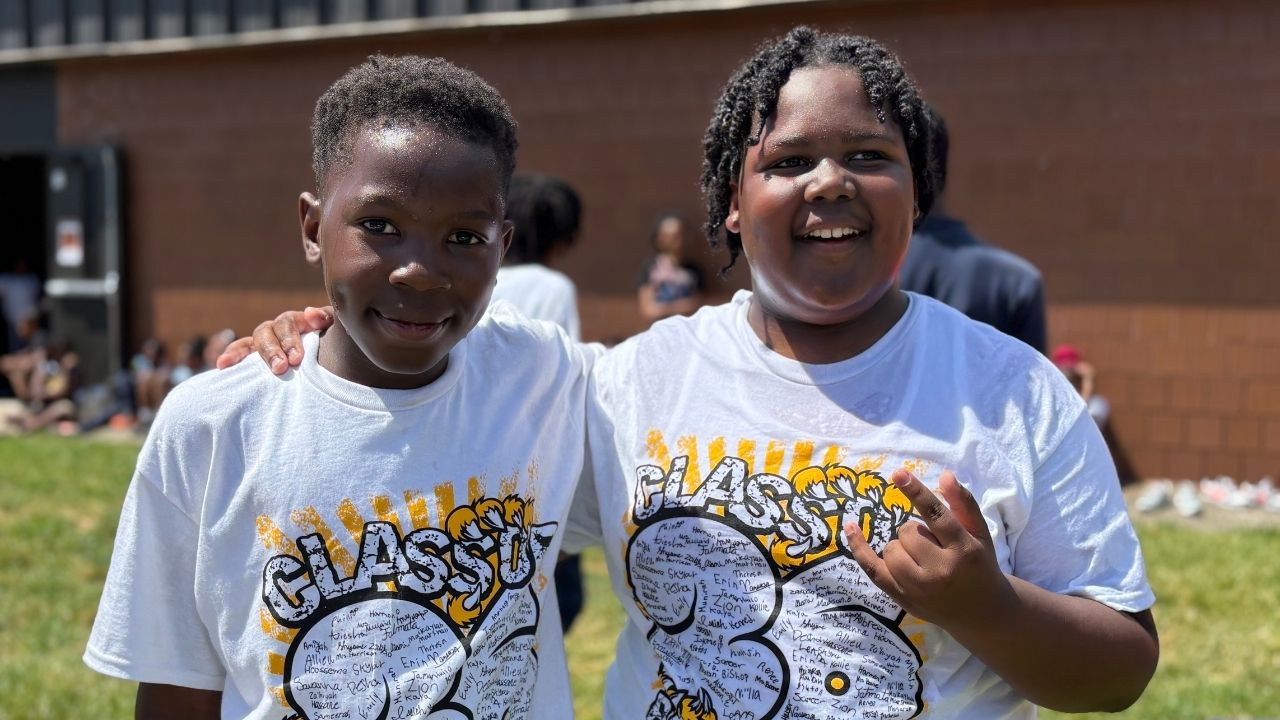 Two smiling students standing side-by-side outdoors wearing matching white 