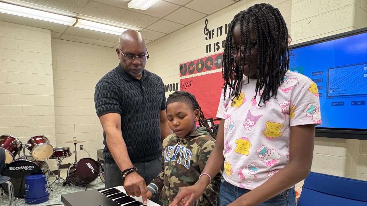 An adult instructor guiding two students playing an electronic keyboard in a music classroom equipped with a drum kit and digital display board.