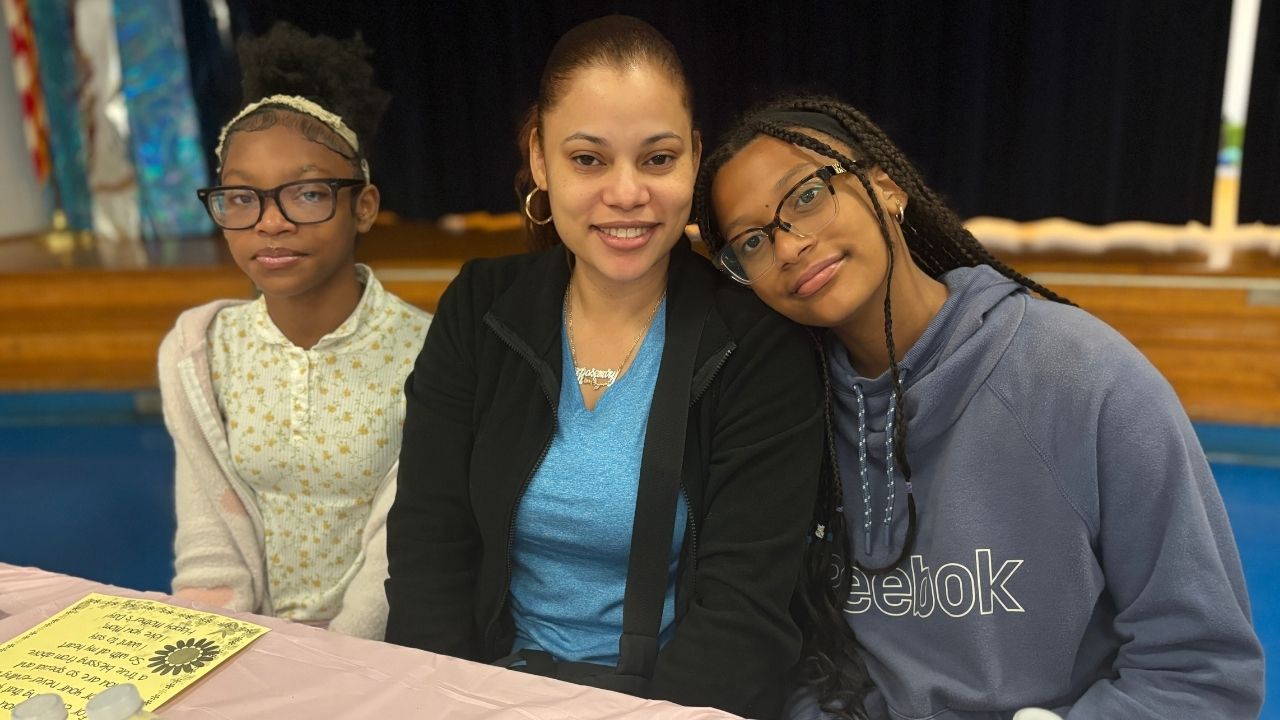 A woman sitting at a table flanked by two young girls, with the girl on the right wearing glasses and a blue Reebok hoodie leaning her head affectionately against her shoulder.