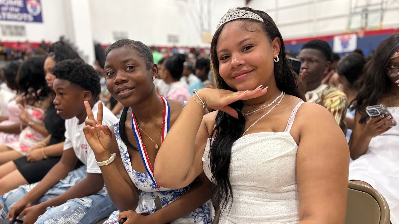 Two smiling students seated together in an auditorium during a formal ceremony; the student on the left flash a peace sign while wearing a blue-and-white patterned dress with a medal, and the student on the right wears a tiara and a sparkly white dress.