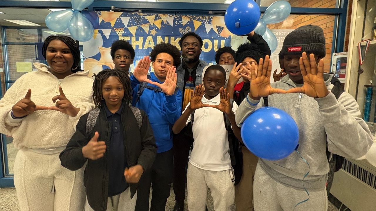 A group of students and an adult posing together in a school hallway with blue and light blue balloons in front of a 