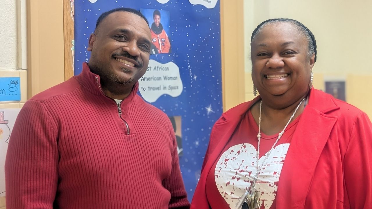 A smiling man in a maroon quarter-zip sweater and a smiling woman in a red blazer and red heart shirt standing side by side in a hallway in front of a bulletin board dedicated to Mae Jemison.