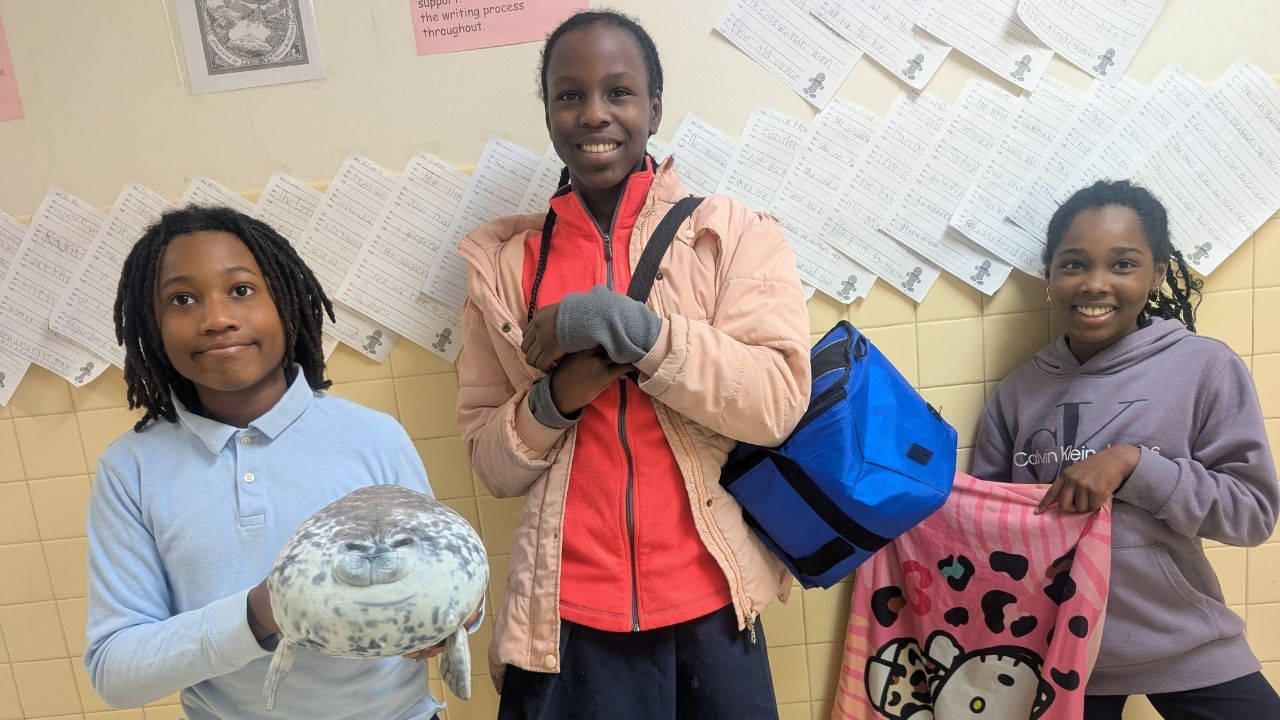 Three smiling students posing together in front of a classroom display wall. The student on the left in a light blue polo holds a round seal plushie, the middle student wears a peach jacket and blue skirt carrying a blue bag, and the student on the right in a gray hoodie holds up a pink Hello Kitty towel.