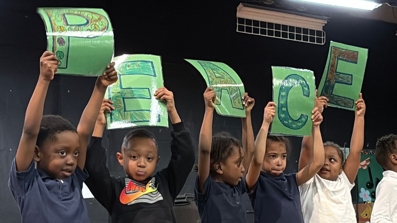 Five young students standing in a row on a stage holding up green signs that spell out the word 