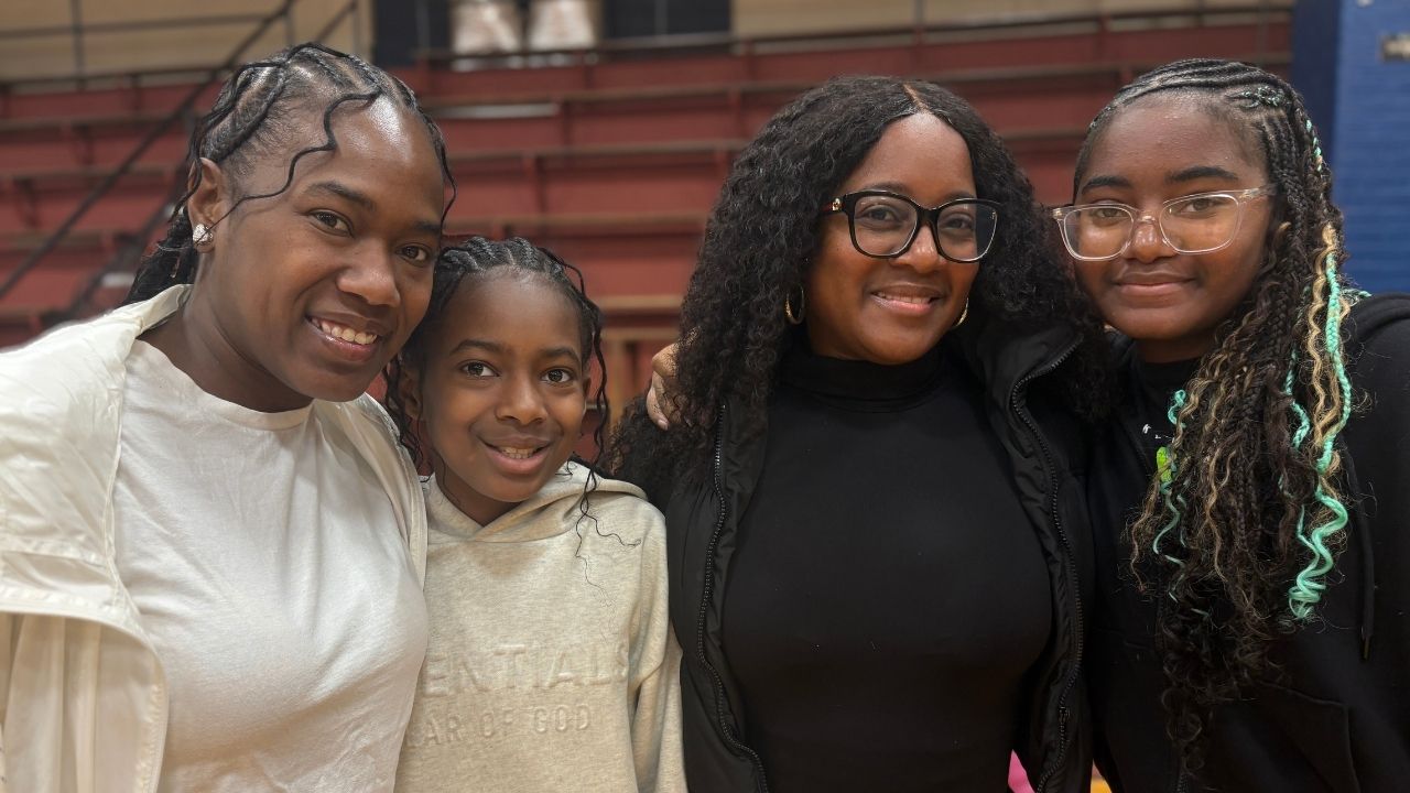 Four smiling people posing together indoors with gym bleachers visible in the background. On the left, a woman in a white jacket stands next to a young girl wearing a beige 