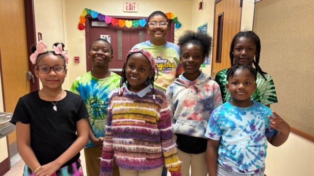 Seven students wearing colorful tie-dye shirts, patterned sweaters, and bright outfits standing together and smiling in a school hallway adorned with colorful heart decorations.