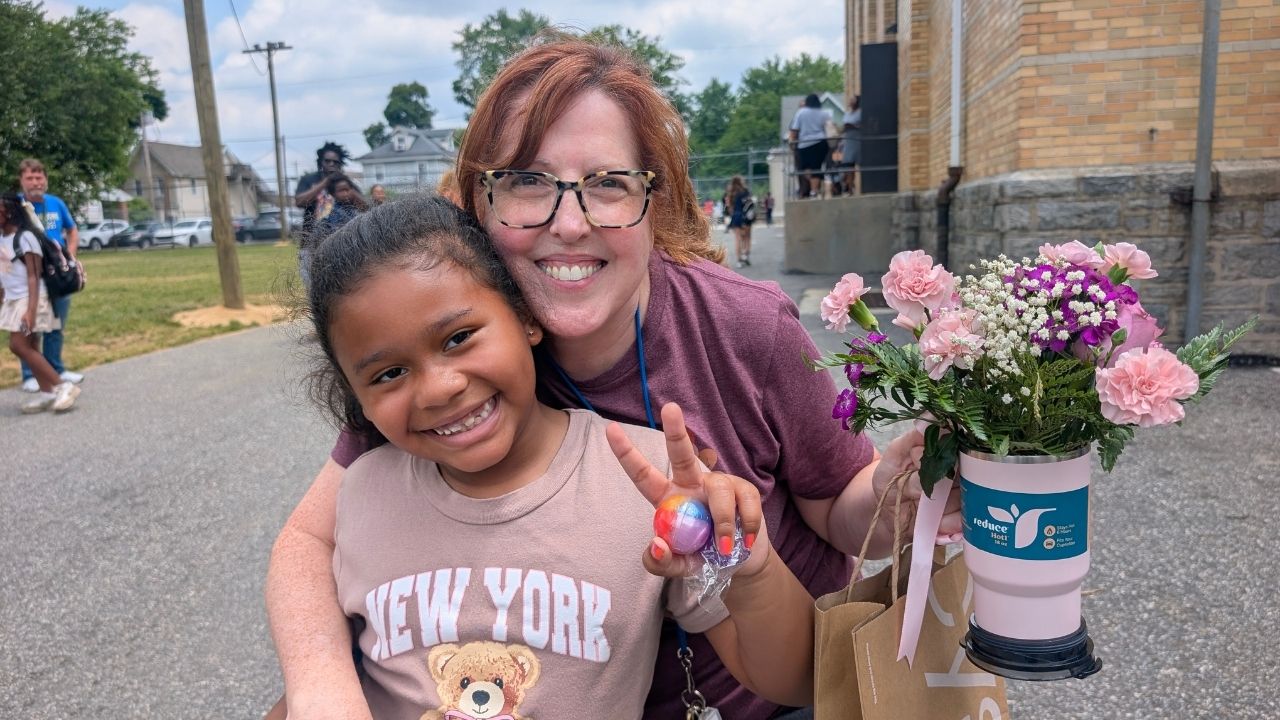 A woman with glasses and maroon shirt hugging a young student wearing a teddy bear shirt outdoors. The girl holds up a peace sign, and the woman holds a gift bag containing a pink travel mug filled with pink and purple flowers.