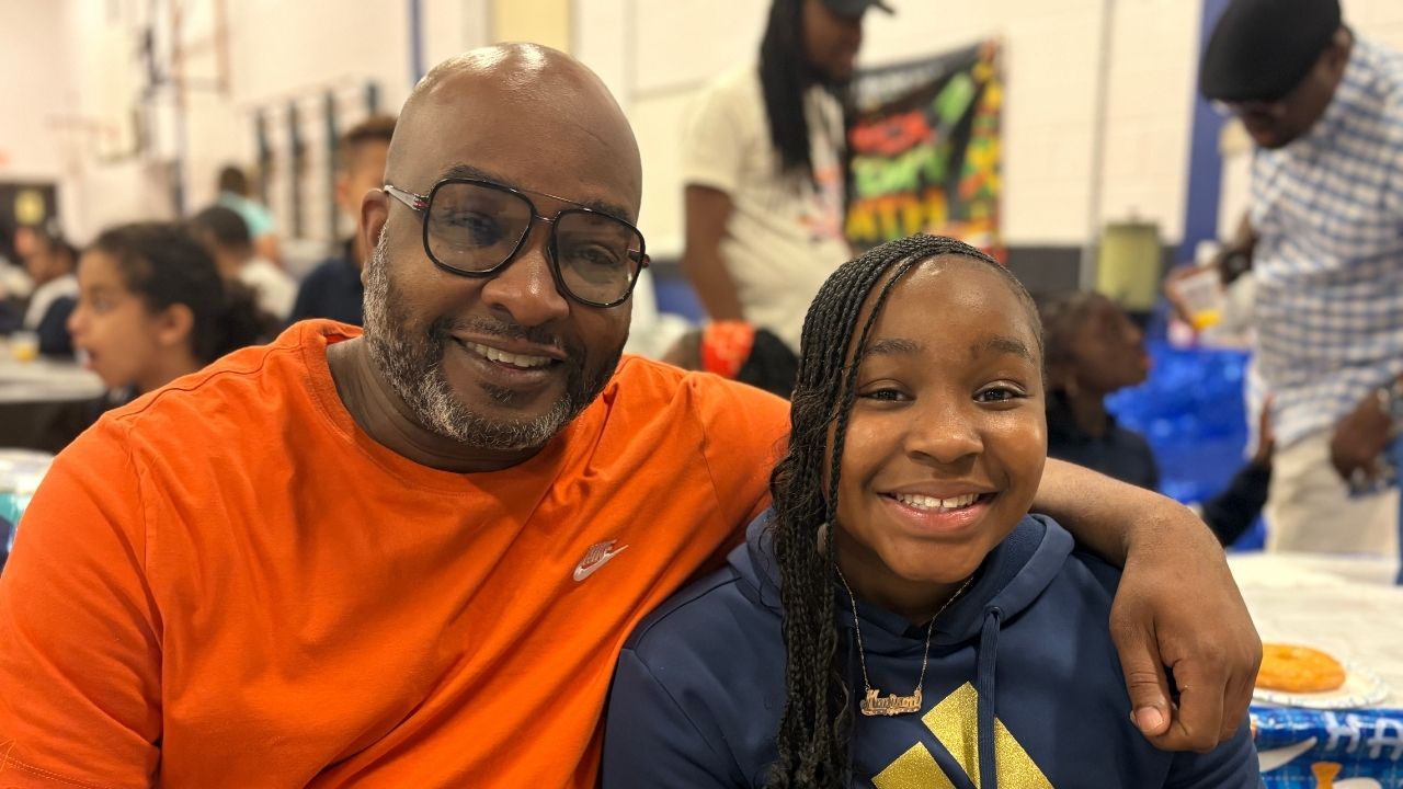 A smiling man wearing black-rimmed glasses and an orange shirt with his arm wrapped around a young girl with braided hair wearing a blue hoodie. They are seated together at a table during an event in a school gymnasium.