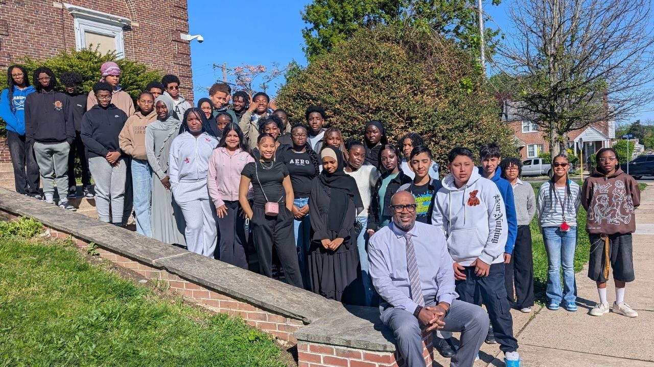 A large group of students gather outdoors on a sunny day along a brick walkway, with a man in a tie and dress clothes seated at the front of the group on a brick wall outside a school building.