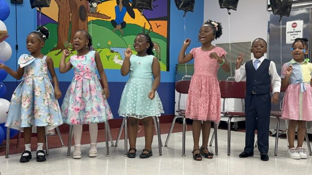A group of six young children five girls in colorful dresses and one boy in a navy blue suit vest stand in a line on a stage, singing or performing. They are positioned in front of a colorful wall mural depicting a forest scene.