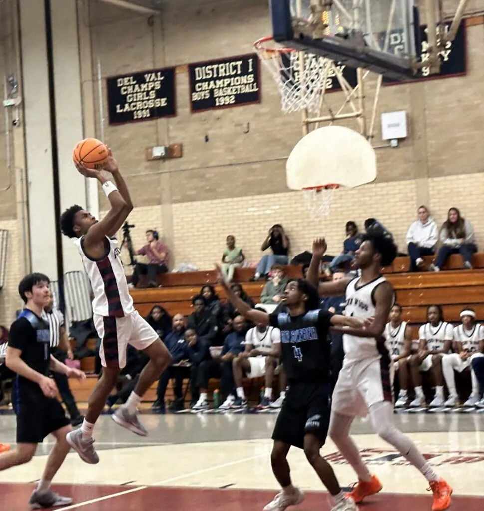 A high school basketball player in a white and maroon Penn Wood jersey jumps for a mid-air shot during a game. He is surrounded by opposing players in black jerseys, with a crowded gymnasium and championship banners in the background.