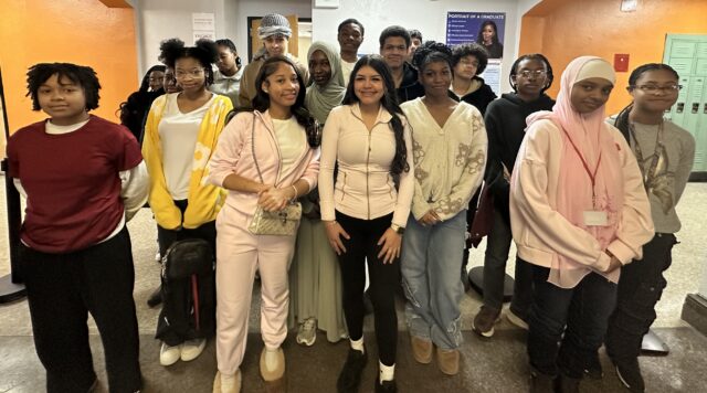 A diverse group of high school students stands together in a school hallway for a group photo. They are wearing a variety of casual clothing, including sweatshirts, cardigans, and head coverings.