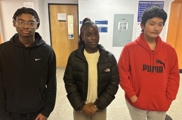 A medium shot of three teenage students, two boys and one girl, standing side-by-side in a school hallway. They are dressed in casual outerwear and looking directly at the camera.