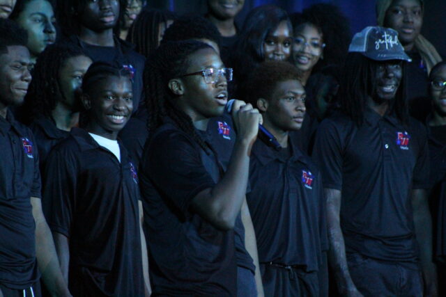 A diverse group of students is gathered on stage, dressed in matching black polo shirts featuring the school's music program logo. A student with long locs and glasses stands at the center, holding a microphone and singing. The students surrounding the soloist are smiling and engaged, appearing to provide backing vocals or support. The lighting is dramatic and cool-toned, typical of an indoor auditorium performance.