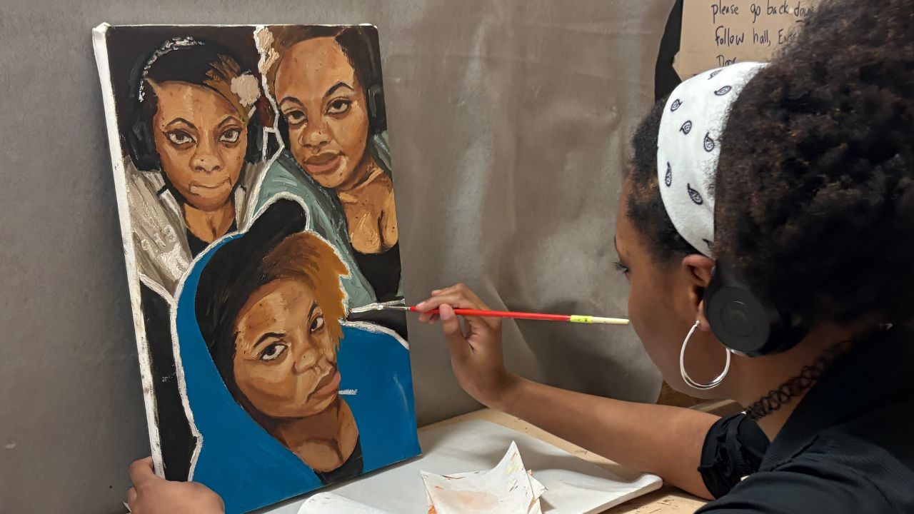 An artist wearing a white patterned headband and headphones works on a canvas painting featuring three expressive portraits of Black women. The artist holds a red paintbrush, focusing on the details of the portrait in the foreground.