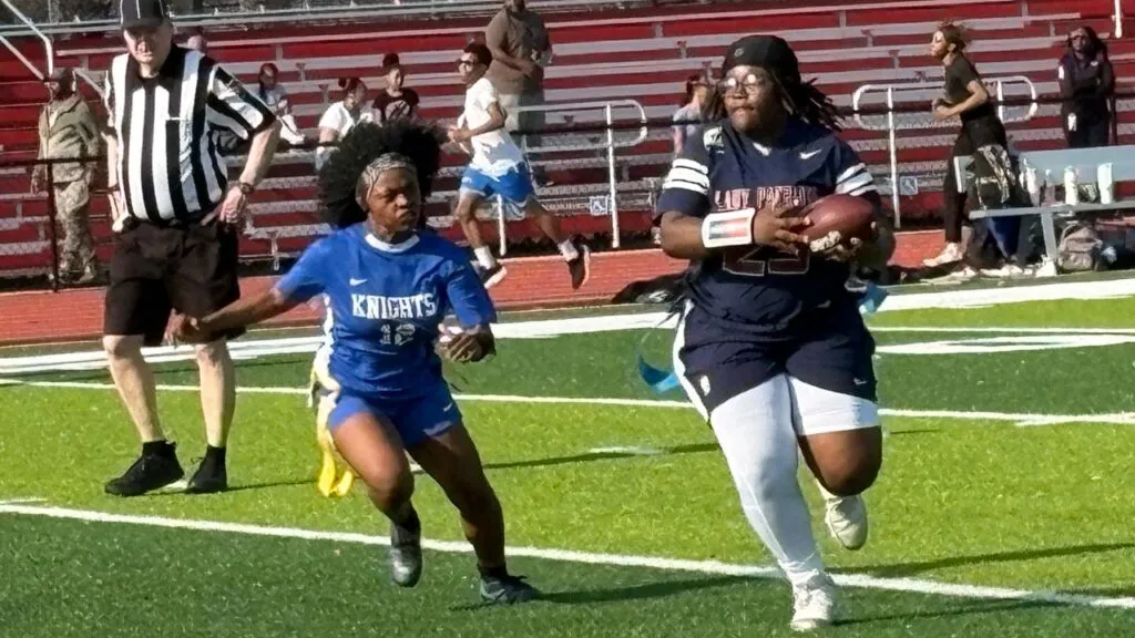 An action shot from a girls' flag football game on a sunny day. In the foreground, a player in a dark blue 