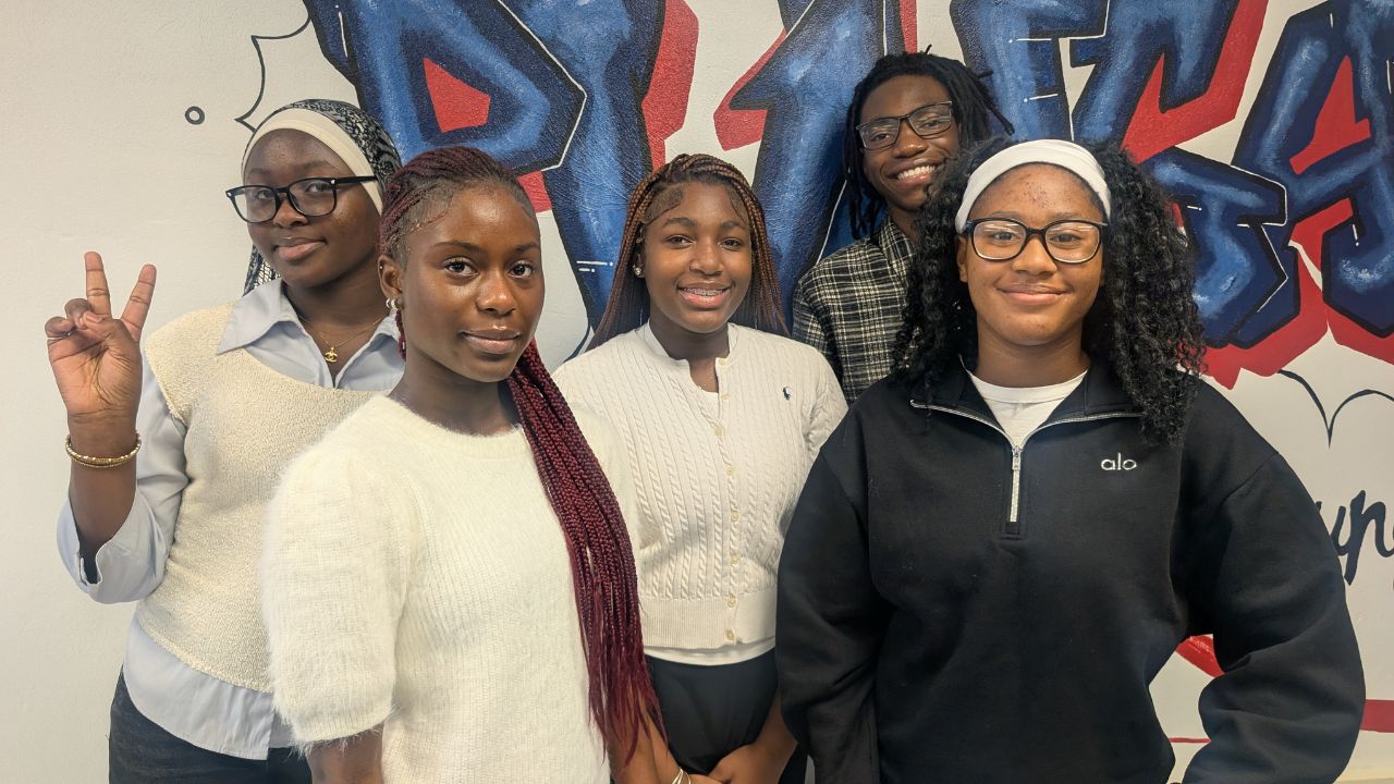 5 students standing smiling in front of a graphic art wall
