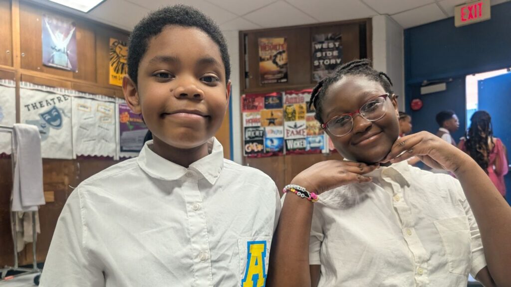 Two smiling children, a boy and a girl, stand next to each other indoors. The boy on the left wears a white button-down shirt with a blue and yellow letter 