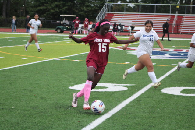 A Penn Wood soccer player wearing jersey number 15 dribbles a soccer ball down an outdoor turf field while a Norristown defender wearing jersey number 27 pursues.